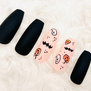 May include: Five long, black coffin-shaped fake nails with a matte finish. Two of the nails have a light pink base with black and orange Halloween-themed designs including ghosts, bats, pumpkins, and candy corn.