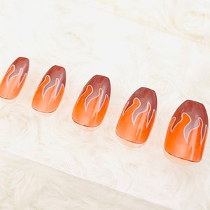 May include: Five oval-shaped false nails with a flame design. The nails transition from a light orange base to a dark brown tip, with white and orange flame details. The nails are arranged on a clear surface.
