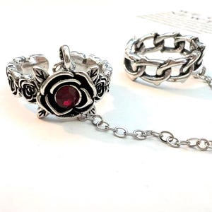 May include: Two-piece silver-toned ring set. One ring features a rose design with a red gemstone center and a chain connecting it to a second ring with a chain link design. The rings are on a white surface.