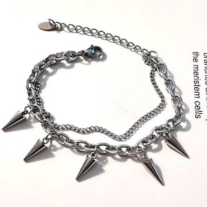 May include: A silver-coloured, layered bracelet featuring multiple chain links and cone-shaped spikes. The bracelet has an adjustable chain. The jewellery is displayed on a white background.