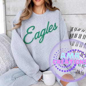 Könnte beinhalten: Ein hellgraues Sweatshirt mit Rundhalsausschnitt und dem Wort Eagles, das in grüner Schreibschrift auf die Brust gestickt ist. Das Kleidungsstück hat lange Ärmel sowie gerippte Bündchen an Ausschnitt, Ärmeln und Saum. Ein AMR CREATIONS Logo ist zu sehen.