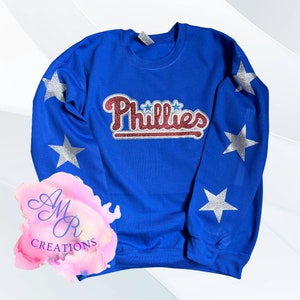 Könnte beinhalten: Ein königsblauer Pullover mit dem Wort "Phillies" in rotem und weißem Glitzer auf der Vorderseite. Die Ärmel haben silberne Glitzersterne.