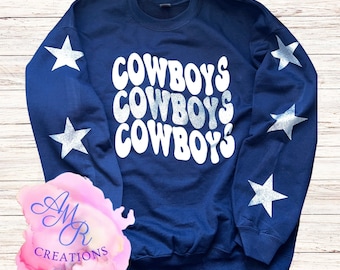Op de afbeelding: Navyblauwe trui met de woorden "COWBOYS" drie keer herhaald in een retro-lettertype. De trui heeft zilveren glittersterren op de mouwen.