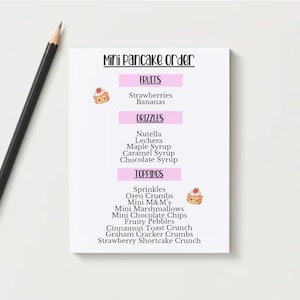 May include: A white notepad with the text "Mini Pancake Order" and a list of fruits, drizzles, and toppings. A black pencil with a gold ferrule lies to the left of the notepad. The notepad has pink accents and cartoon pancake illustrations.