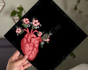 Może przedstawiać: Czarna czapka absolwenta z szczegółową ilustracją ludzkiego serca ozdobionego różowymi kwiatami i zielonymi liśćmi. Serce jest czerwone, a kwiaty mają białe akcenty. Tekst "Kim's Custom Caps" jest wydrukowany po przekątnej na czapce.