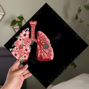 Könnte beinhalten: Eine schwarze Abschluss-Kappe mit einer roten und weißen Illustration einer menschlichen Lunge mit Blumen. Die Lunge ist stilisiert mit sichtbaren Adern und Arterien.