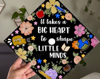 May include: Black graduation cap decorated with colorful flowers, a school bus, a globe, a book, a paint palette, and a pencil. The text "It takes a BIG HEART to shape LITTLE MINDS" is written in white on the cap.