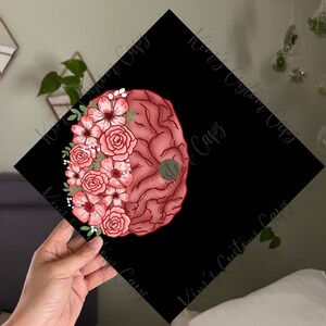 May include: A black graduation cap with a red brain and pink flowers. The brain is surrounded by a wreath of pink flowers with green leaves.
