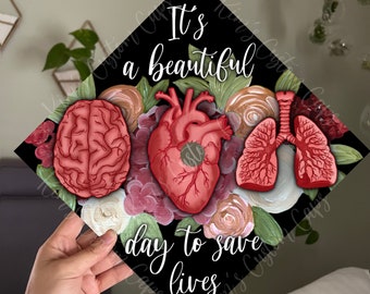 May include: Black graduation cap with a floral design and anatomical illustrations of a brain, heart, and lungs. The text "It's a beautiful day to save lives" is printed on the cap.