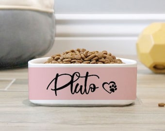 May include: A pink and white pet bowl filled with dry dog food. The bowl is personalized with the name "Pluto" in black script, a heart, and a paw print. A yellow toy and a gray bowl are in the background.