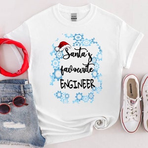 Op de afbeelding: Wit T-shirt met de tekst "Santa's favourite ENGINEER" in zwart schrift, omringd door blauwe tandwielen en een kerstmuts. Het shirt is gestyled met een spijkerbroek, rode zonnebril, rode bandana en witte sneakers.