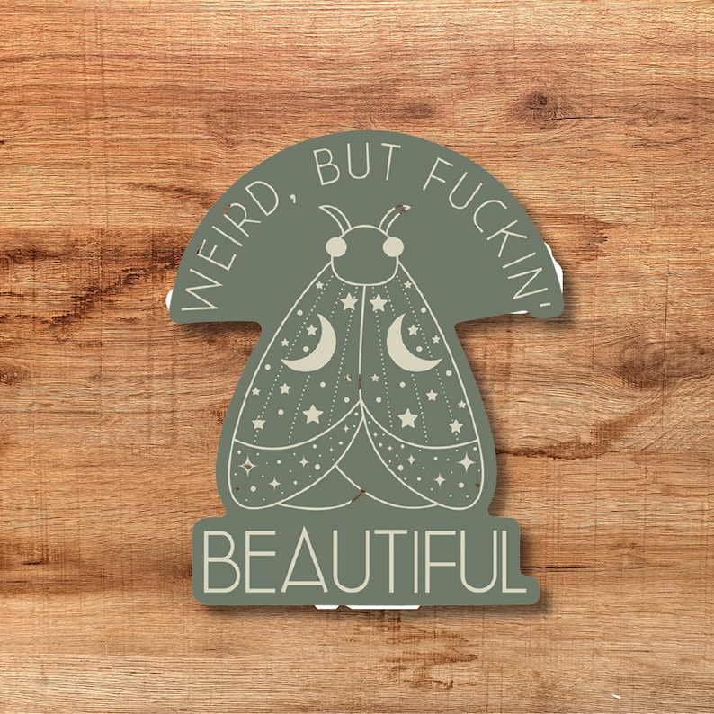 May include: A sticker with a moth design in shades of green and cream, featuring crescent moons and stars. The text reads "WEIRD, BUT FUCKIN' BEAUTIFUL" in a circular and stacked layout. The sticker is set against a wooden background.