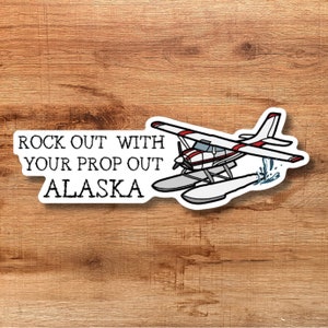 May include: A white sticker featuring a seaplane with red and white accents, and the text "ROCK OUT WITH YOUR PROP OUT ALASKA". The sticker is on a wooden background.