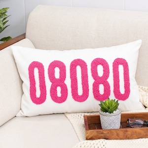 May include: A white couch with a white throw blanket and a pillow with the number 08080 in pink. A small potted plant and sunglasses are on a wooden tray on the couch.