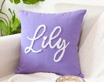 May include: A purple throw pillow with the name "Lily" embroidered in white on the front. The pillow is sitting on a white couch with a white throw blanket and a wicker candle holder.