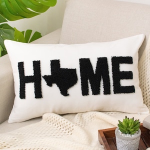 May include: White throw pillow with black embroidered text that reads "HOME" with the state of Texas in the middle of the word. The pillow is on a white couch with a white throw blanket and a small potted plant.
