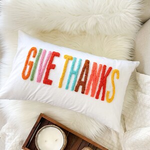Peut inclure: Un coussin blanc avec les mots "GIVE THANKS" brod&eacute;s en arc-en-ciel. Le coussin est sur une couverture en fausse fourrure blanche avec un plateau en bois contenant une bougie et des boules d&eacute;coratives.