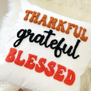 Könnte beinhalten: Weißes Zierkissen mit den gestickten Wörtern "THANKFUL", "grateful" und "BLESSED" in Orange und Schwarz. Das Kissen liegt auf einer weißen, flauschigen Oberfläche, passend für die Inneneinrichtung.