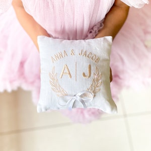 May include: White pillow with gold embroidery that says "Anna & Jacob" with the initials "AJ" in a wreath. The pillow has a white satin ribbon tied in a bow.