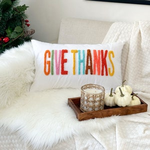 Peut inclure: Un coussin blanc avec les mots "GIVE THANKS" brod&eacute;s en fil color&eacute;. Le coussin est sur une couverture en fausse fourrure blanche et une couverture blanche &agrave; motif tiss&eacute;. Un plateau en bois avec une bougie blanche et des citrouilles blanches est sur la couverture.