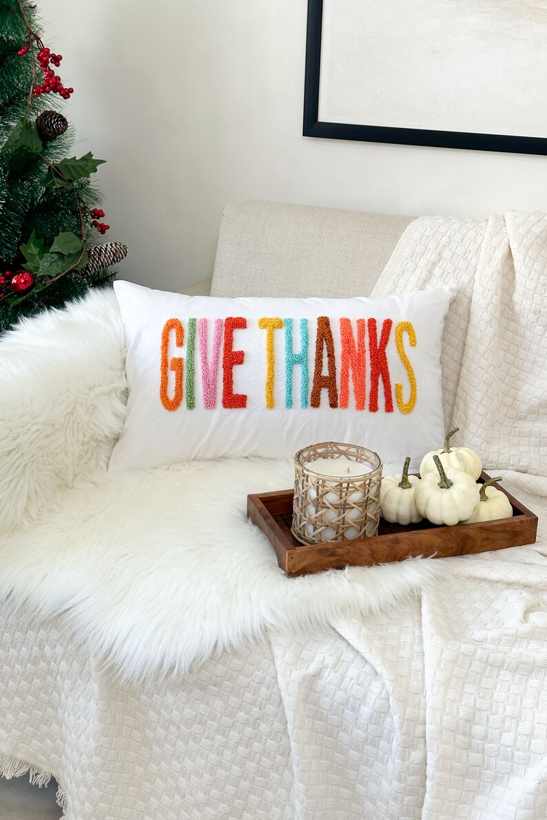 Peut inclure: Un coussin blanc avec les mots "GIVE THANKS" brod&eacute;s en fil color&eacute;. Le coussin est sur une couverture en fausse fourrure blanche et une couverture blanche &agrave; motif tiss&eacute;. Un plateau en bois avec une bougie blanche et des citrouilles blanches est sur la couverture.