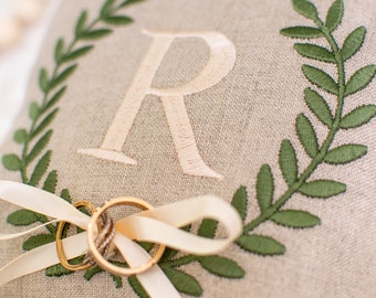 May include: A beige linen ring pillow embroidered with a green laurel wreath and the letter "R" in a light beige thread. Two gold wedding rings are tied to the pillow with a cream-colored ribbon. The pillow is a wedding accessory.