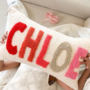 Könnte beinhalten: Ein weißes Kissen mit dem Namen "CHLOE" in bunter Wolle. Die Buchstaben bestehen aus Pompons in den Farben Rot, Orange, Pink und Grau.