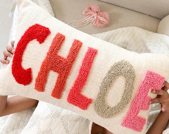 Peut inclure: Un oreiller blanc avec le nom "CHLOE" écrit en fil coloré. Les lettres sont faites de pompons dans des tons de rouge, orange, rose et gris.
