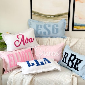 Peut inclure: Six coussins d&eacute;coratifs avec des noms personnalis&eacute;s brod&eacute;s en lettres color&eacute;es. Les coussins sont dispos&eacute;s sur un canap&eacute; blanc avec une couverture blanche.