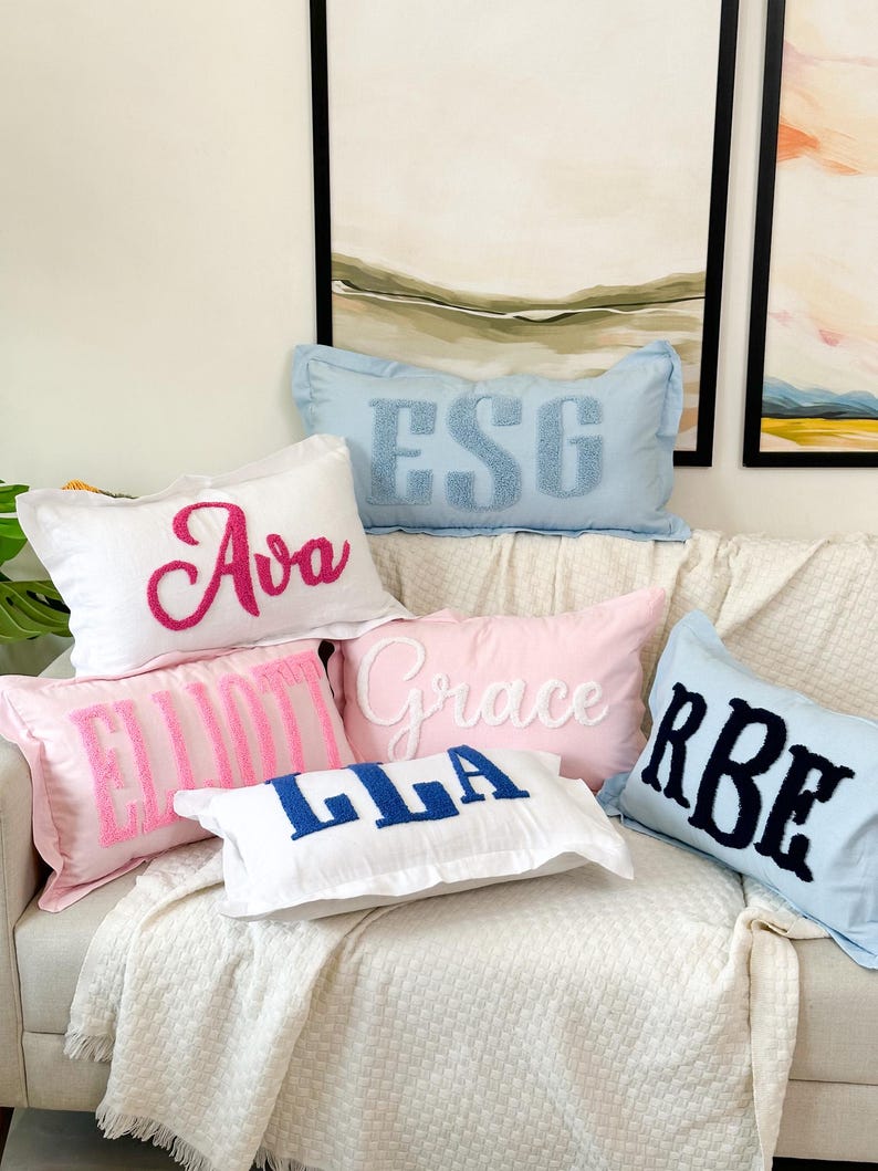 Peut inclure: Six coussins d&eacute;coratifs avec des noms personnalis&eacute;s brod&eacute;s en lettres color&eacute;es. Les coussins sont dispos&eacute;s sur un canap&eacute; blanc avec une couverture blanche.