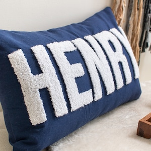 May include: A blue pillow with the name "HENRY" spelled out in white fluffy letters.