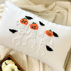 May include: White throw pillow with three embroidered skeletons wearing pumpkin heads. The skeletons are in a line and have black bats flying around them.