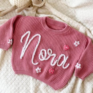 Puede incluir: Un suéter de punto rosa con el nombre "Nora" bordado en escritura blanca. El suéter está decorado con aplicaciones de flores blancas y rosas. Un juguete de oveja de ganchillo está en el fondo.