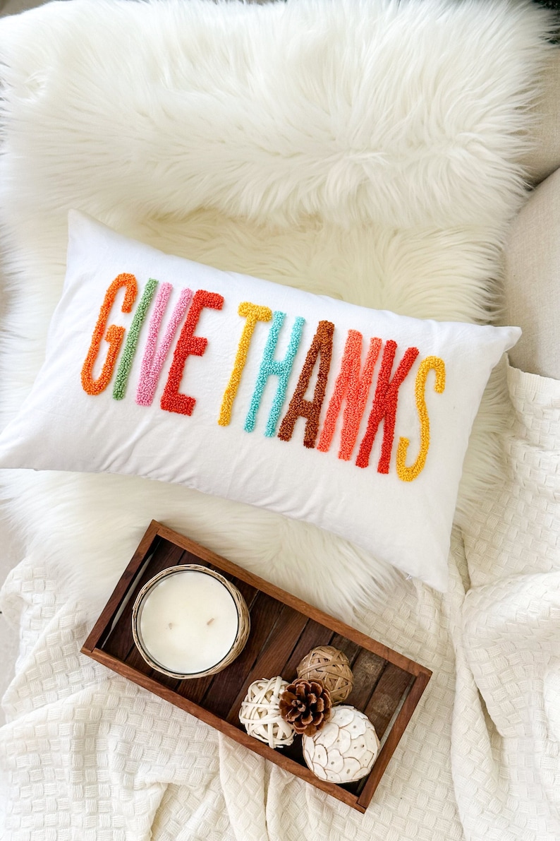 Peut inclure: Un coussin blanc avec les mots "GIVE THANKS" brod&eacute;s en arc-en-ciel. Le coussin est sur une couverture en fausse fourrure blanche avec un plateau en bois contenant une bougie et des boules d&eacute;coratives.