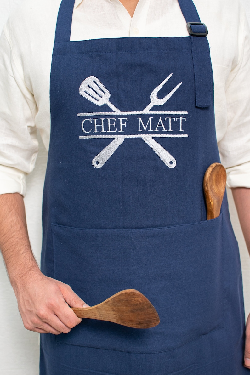Pu&ograve; includere: Un grembiule blu navy con una grande tasca e spalline regolabili. Il grembiule presenta un design ricamato bianco di utensili da cucina incrociati e il testo "CHEF MATT".