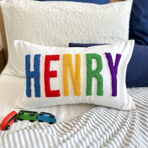 Könnte beinhalten: Ein weißes Kissen mit dem Namen "HENRY", der in Regenbogenfarben gestickt ist. Das Kissen liegt auf einem Bett mit einer weiß-blau gestreiften Bettdecke.