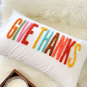 Peut inclure: Un coussin blanc avec les mots "Give Thanks" brod&eacute;s en fil color&eacute;. Les lettres sont dans un sch&eacute;ma de couleurs arc-en-ciel, chaque lettre &eacute;tant d'une couleur diff&eacute;rente.