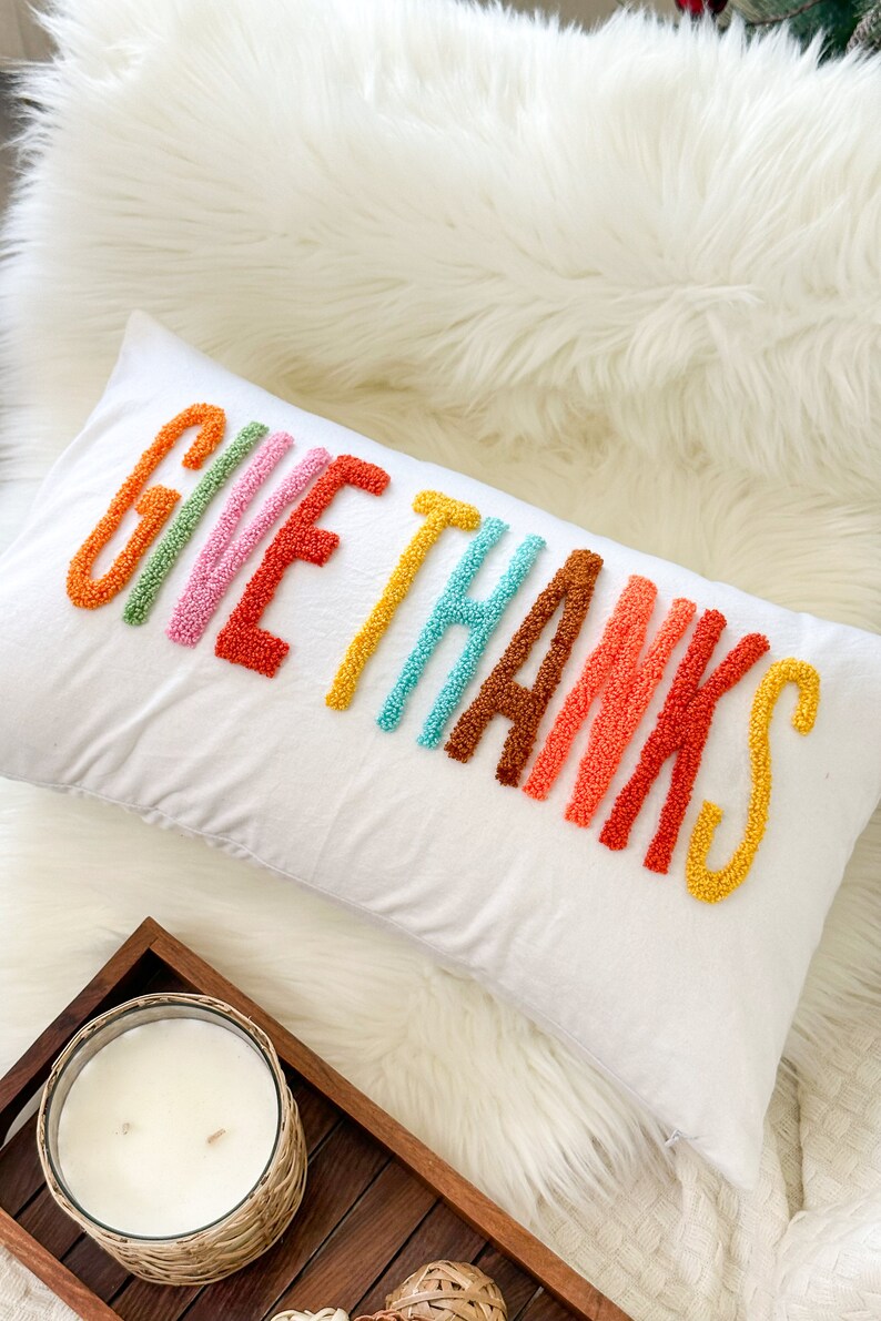 Peut inclure: Un coussin blanc avec les mots "Give Thanks" brod&eacute;s en fil color&eacute;. Les lettres sont dans un sch&eacute;ma de couleurs arc-en-ciel, chaque lettre &eacute;tant d'une couleur diff&eacute;rente.