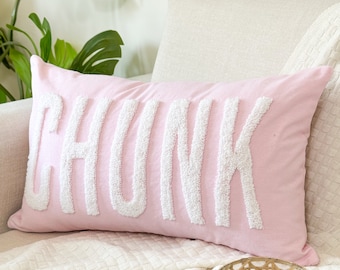 May include: A pink throw pillow with the word "CHUNK" embroidered in white on the front. The pillow is sitting on a white couch with a white blanket and a wicker candle holder.