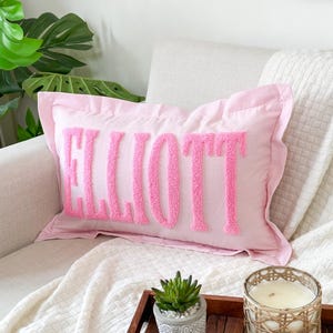 May include: Pink pillow with the name "ELLIOTT" embroidered in pink fluffy letters. The pillow is on a white couch with a white blanket and a wooden tray with a succulent plant and a candle.