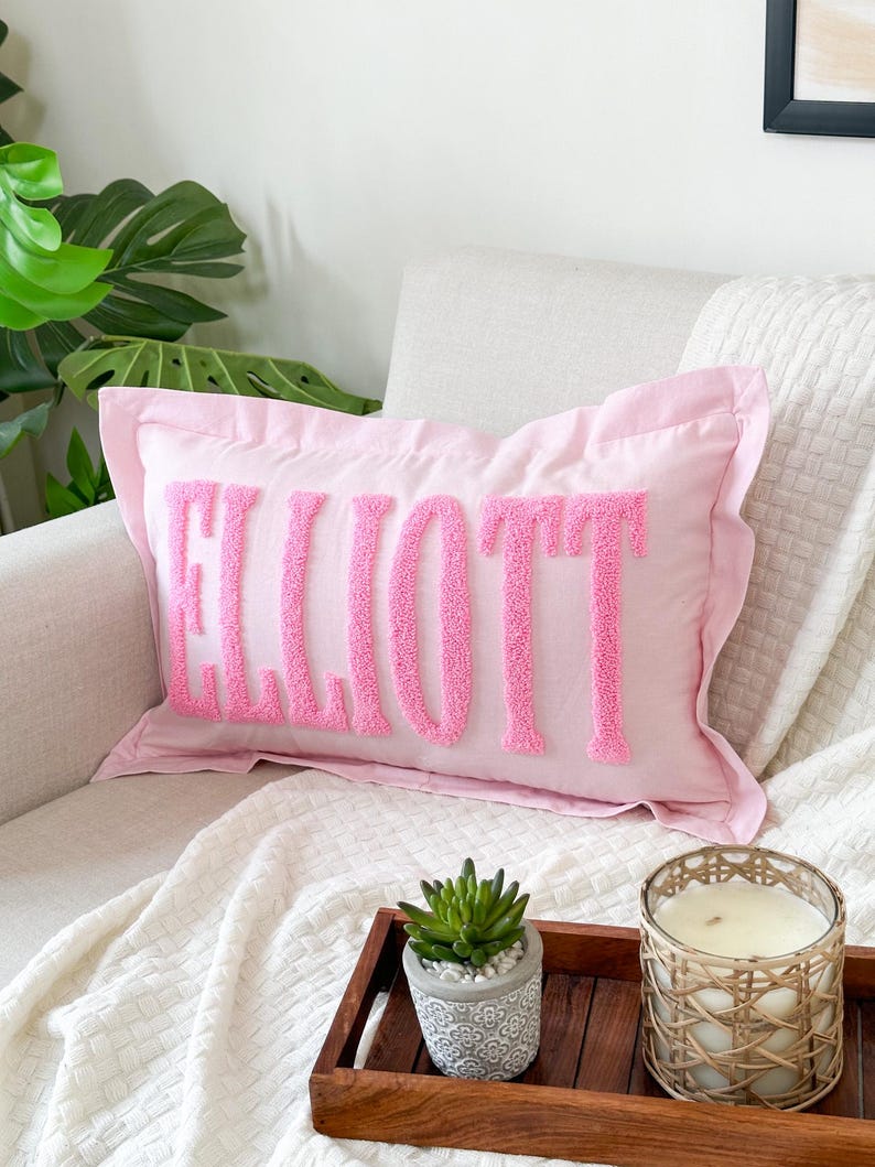 Peut inclure: Un coussin rose avec le nom "ELLIOTT" brod&eacute; en lettres moelleuses roses. Le coussin est sur un canap&eacute; blanc avec une couverture blanche et un plateau en bois avec une plante succulente et une bougie.