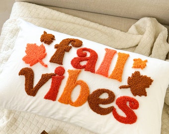 Puede incluir: Una almohada blanca con las palabras "fall vibes" en hilo naranja, rojo y marrón. Las letras están hechas con un hilo esponjoso y texturizado y están rodeadas de pequeñas formas de hojas naranja y marrón.
