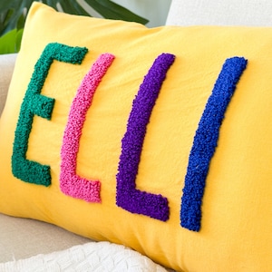 Puede incluir: Una almohada amarilla con el nombre "ELLI" bordado en hilo verde, rosa, morado y azul.