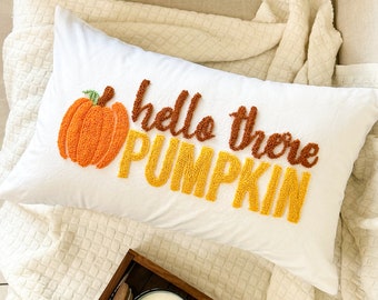 Puede incluir: Una almohada blanca con el texto "hello there pumpkin" en hilo naranja y amarillo. La almohada está sobre una manta de color crema con una bandeja de calabazas blancas y una vela.