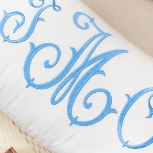 Puede incluir: Una almohada blanca con un monograma bordado en azul. El monograma es un "EOC" estilizado con un adorno decorativo.