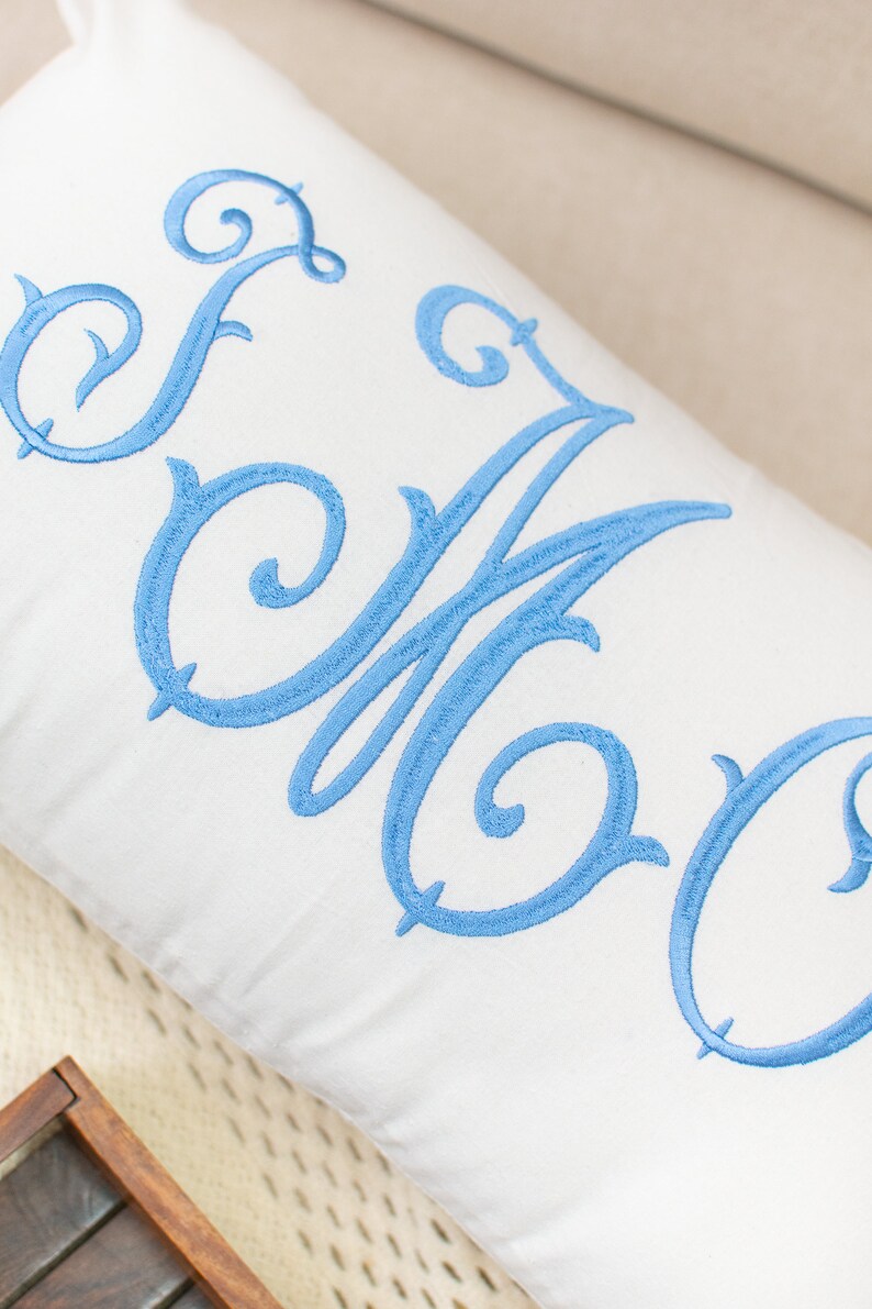 Puede incluir: Una almohada blanca con un monograma bordado en azul. El monograma es un "EOC" estilizado con un adorno decorativo.