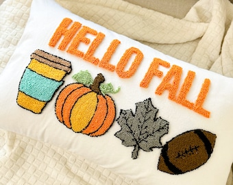 Puede incluir: Cojín rectangular blanco con las palabras "HELLO FALL" en naranja. Presenta una taza de café, una calabaza, una hoja y un balón de fútbol. El diseño está creado con la técnica de aguja mágica, ideal para el otoño.