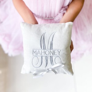 May include: White linen ring pillow with a gray embroidered monogram and the name "MAHONEY" in a script font. The pillow is tied with a white satin ribbon.