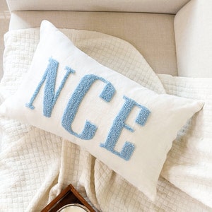 May include: A white throw pillow with the word "NICE" embroidered in light blue on a white background. The pillow is lying on a white blanket with a woven texture.