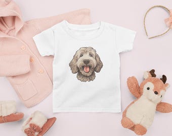 May include: A white toddler t-shirt featuring a detailed illustration of a smiling, light brown dog with its tongue out. The shirt is surrounded by a pink cardigan, a plush deer toy, and other baby accessories.